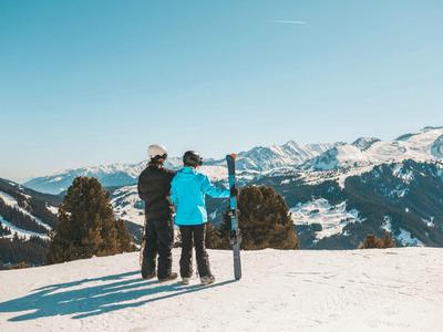Zwei Personen stehen im Schnee, der eine hält ein Snowboard, im Hintergrund Berge und blauer Himmel.