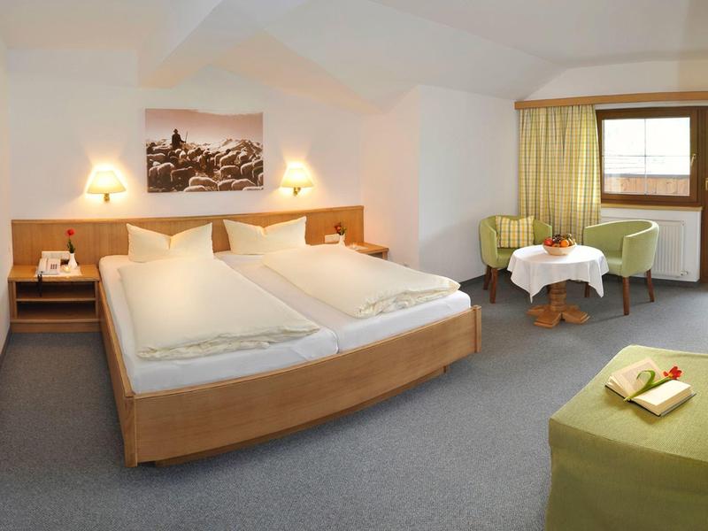 Ein helles Hotelzimmer mit großem Doppelbett, zwei Nachttischen, Sitzbereich und Fenster mit gelben Vorhängen.