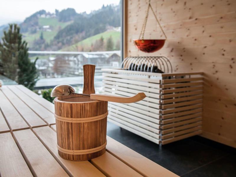 Holzsauna mit Holzbänken, Eimer und Kelle, Blick auf grüne Berge durch Fenster.