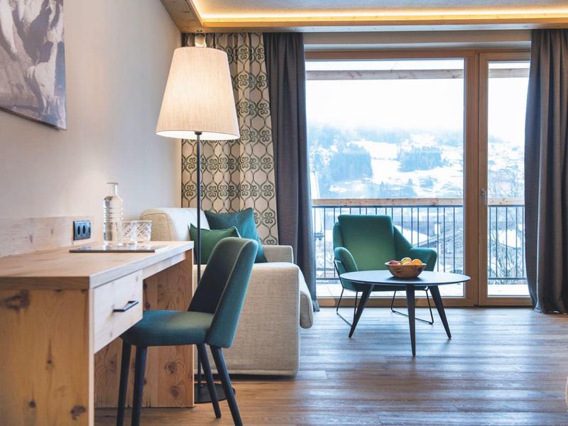 Heller Wohnraum mit Holzboden, Fensterfront, türkisfarbenem Stuhl, Sofa und kleinem Tisch.