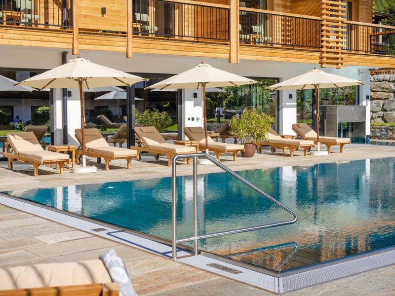 Moderner Außenpool mit Liegestühlen, Sonnenschirmen und Holzterrasse vor einem mehrstöckigen Gebäude.