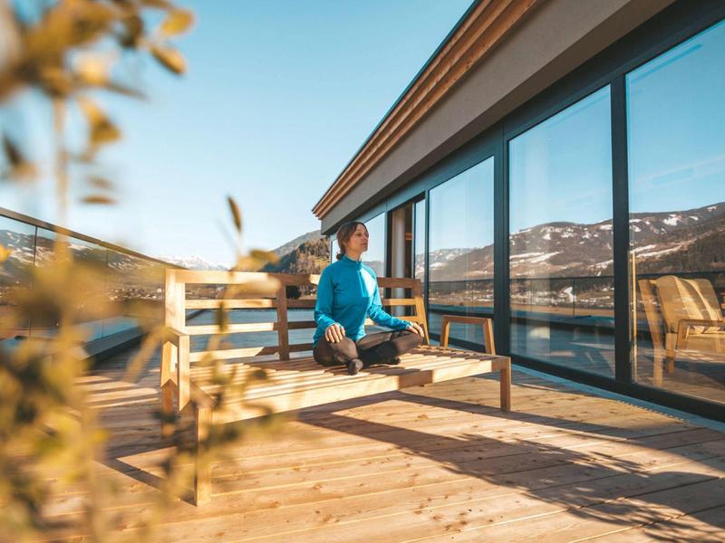 Person meditiert im Schneidersitz auf Terrasse eines modernen Hauses mit Bergblick und Glasfassade.
