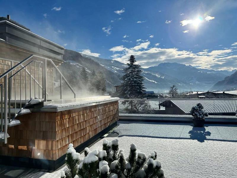 Winterlandschaft mit schneebedeckten Dächern, dampfendem Holzpool und Bergen im Hintergrund.