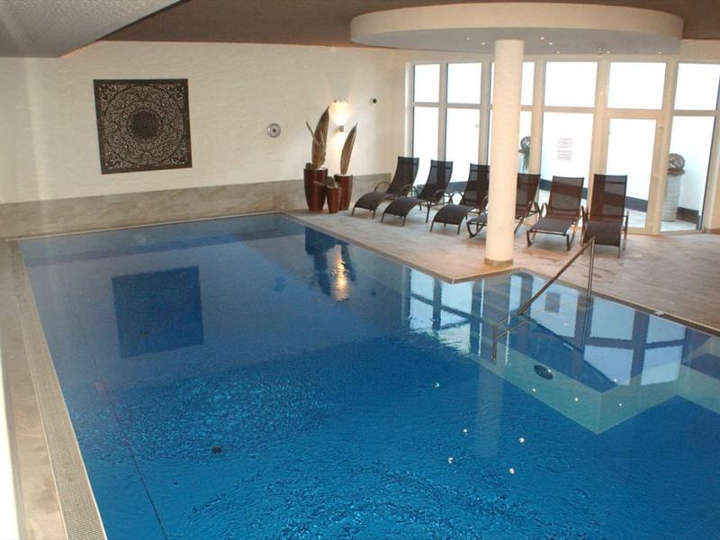 Hell beleuchteter Raum mit großem, blauem Hallenbad und mehreren Liegestühlen am Fenster.