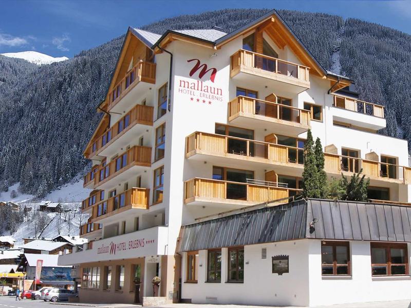 Mehrstöckiges Hotel mit Holzbalustraden vor schneebedecktem Bergpanorama und blauem Himmel.