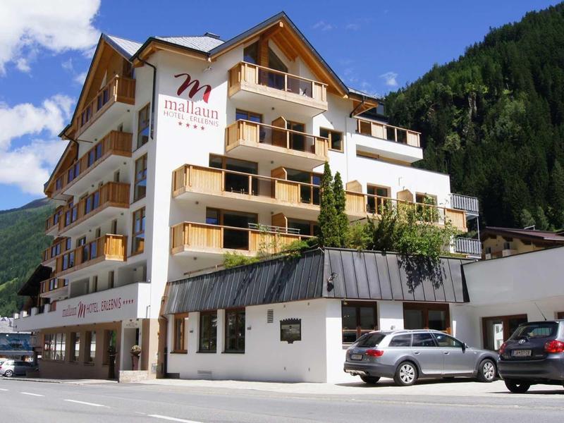 Mehrstöckiges Hotelgebäude mit Holzbalkonen und Autos davor, umgeben von Bergen und blauem Himmel.