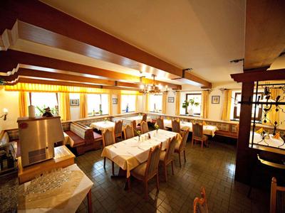 Ein heller Gastraum mit Holzbalken, weißen Tischen und gelben Vorhängen an den Fenstern.