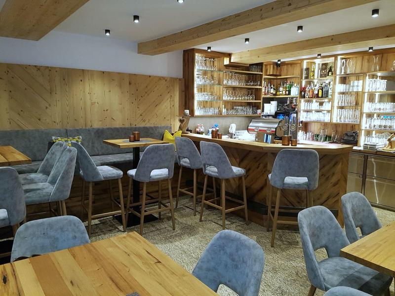 Moderne Bar mit Holzvertäfelung, grauen Stühlen und Theke, hellem Holzboden und Regalen mit Gläsern.