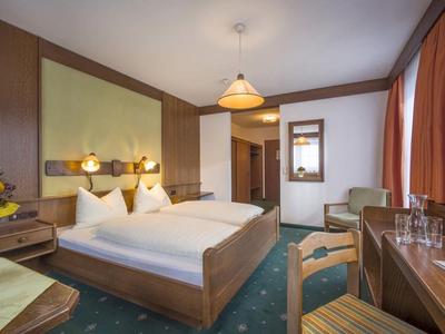 Hell eingerichtetes Hotelzimmer mit Doppelbett, Holzmöbeln, grünem Teppich und orangen Vorhängen.