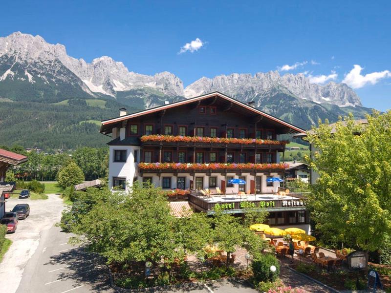 Gasthaus mit Holzterrasse und Garten, im Hintergrund hohe Berge unter blauem Himmel.