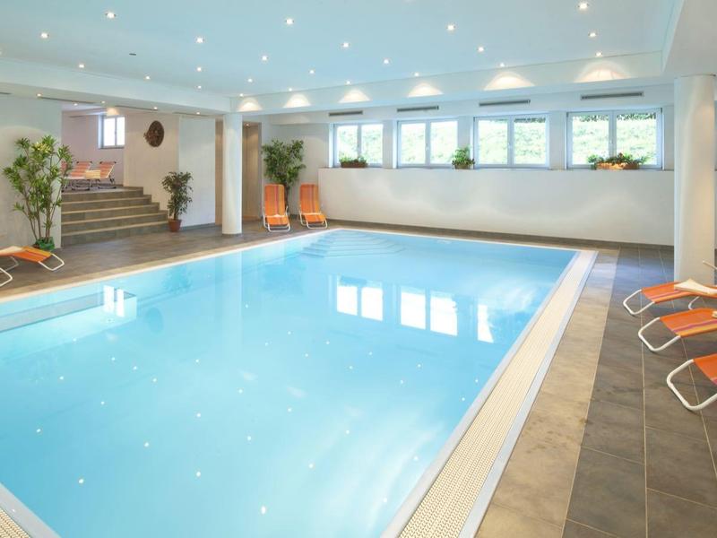 Heller Hallenbadbereich mit blauem Wasser, orangen Liegestühlen und Pflanzen in moderner Umgebung.