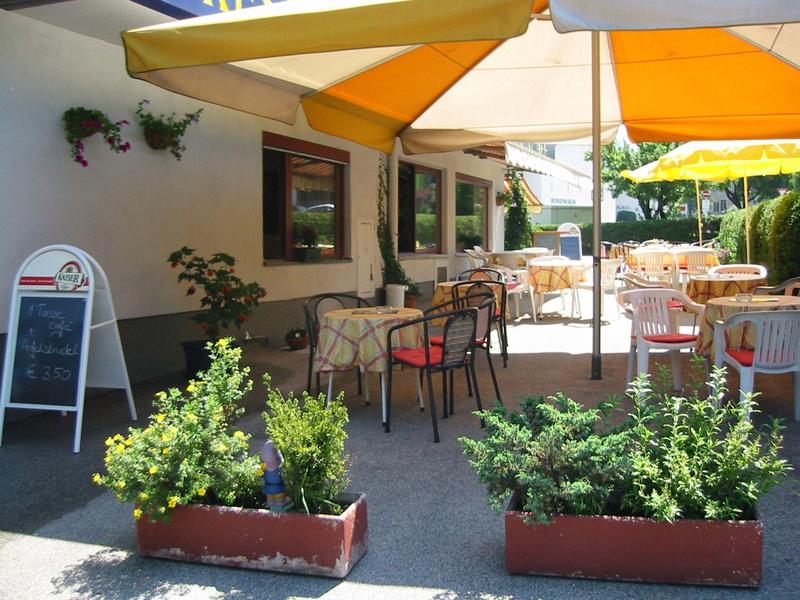 Sonniger Cafébereich mit Stühlen, Tischen, Pflanzen und gelben Markisen an Hauswand.