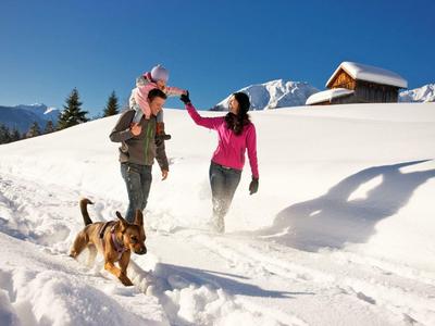 Winterlandschaft mit schneebedecktem Weg, zwei Menschen beim Spielen und ein laufender Hund.
