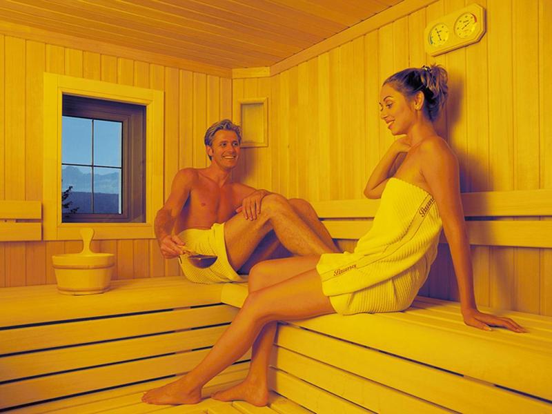 Zwei Frauen entspannt in hellem Sauna-Raum mit Holzwänden und Fenster bei gedämpftem Licht.