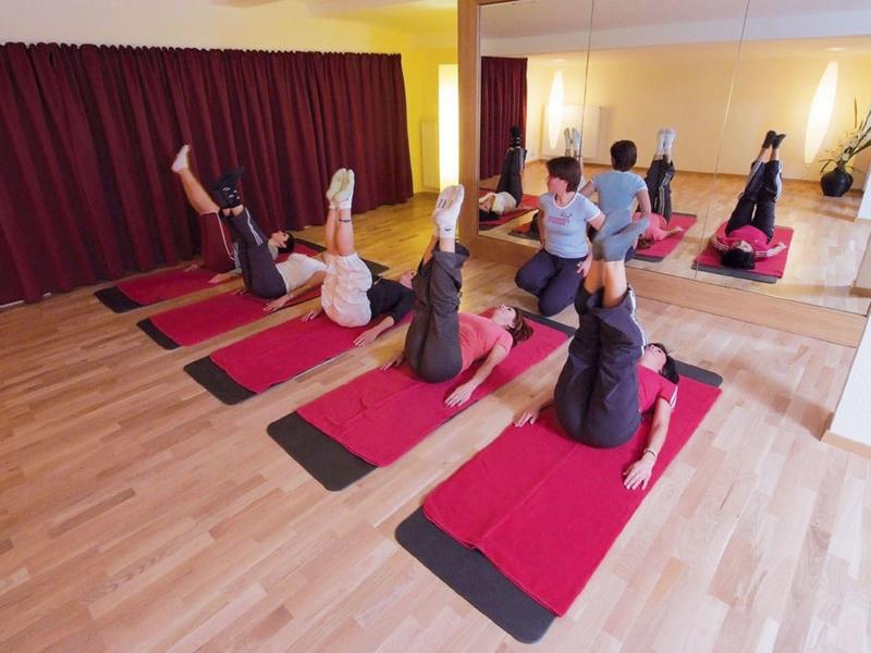 Vier Personen auf roten Yogamatten machen Übungen in hellem Raum mit Spiegelwand und roten Vorhängen.