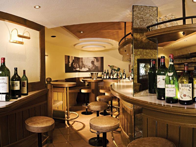 Gemütliche Bar mit Holztheke, runden Hockern, Weinflaschen und warmem Licht in rustikalem Stil.