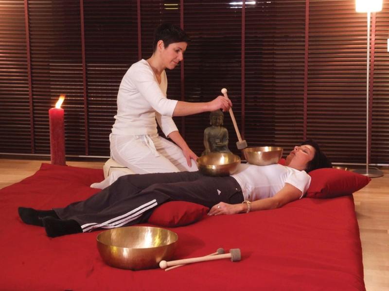 Frau legt Klangschale auf Brust eines Mannes, beide auf roter Matte, ruhige und entspannte Atmosphäre.