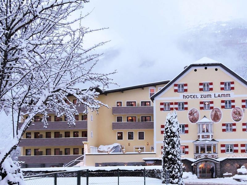 Schneebedecktes Hotel mit traditionellen rot-weißen Fensterläden und schneebedeckten Bäumen davor.
