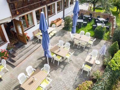 Strefa zewnętrzna hotelu z stolikami, krzesłami i parasolami na utwardzonym terenie obok ogrodu.