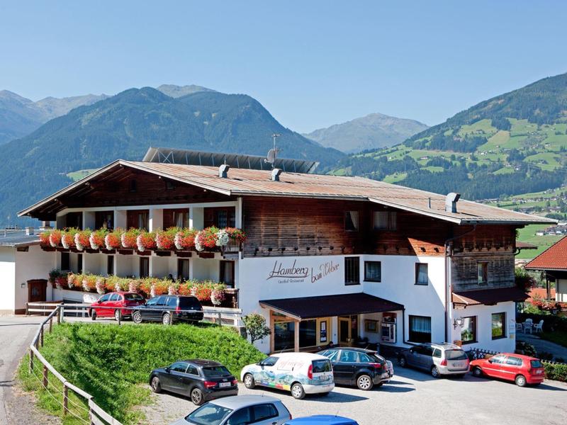 Traditionelles Alpenhaus mit Blumenbalkonen, Autos davor, umgeben von grünen Bergen.