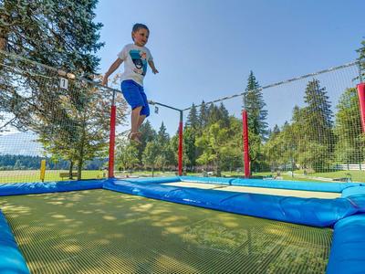Kind springt auf blauem Trampolin im Freien, umgeben von grünen Bäumen und strahlend blauem Himmel.