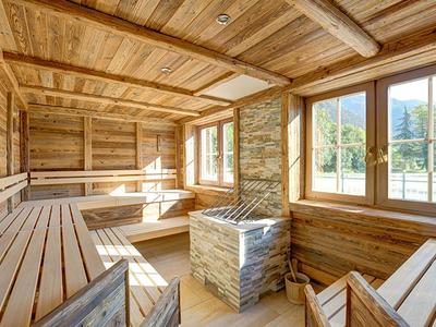 Hell beleuchteter Holz-Saunaraum mit Holzbänken, großen Fenstern und einem Steinkamin im Zentrum.