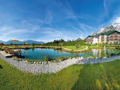 Weitläufige grüne Wiese mit kleinem Teich, Bäumen und großem Hotelgebäude vor Bergkulisse bei blauem Himmel.
