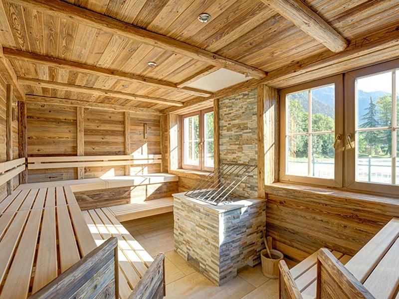 Hell beleuchteter Holz-Saunaraum mit Holzbänken, großen Fenstern und einem Steinkamin im Zentrum.