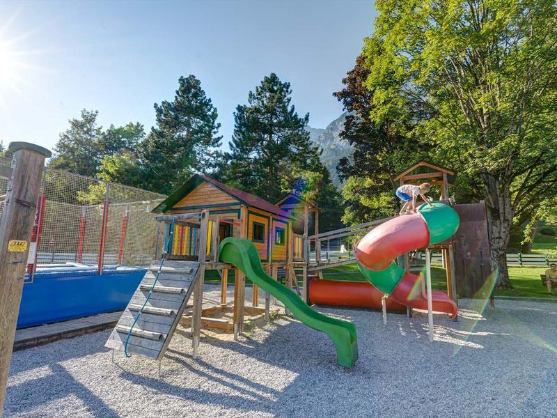 Ein bunter Spielplatz mit Rutsche, Klettergerüst und Sandfläche umgeben von Bäumen und Sonnenschein.