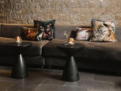 Przytulny kącik wypoczynkowy z ciemną sofą, ozdobnymi poduszkami i małymi czarnymi stolikami przy ścianie z cegieł.