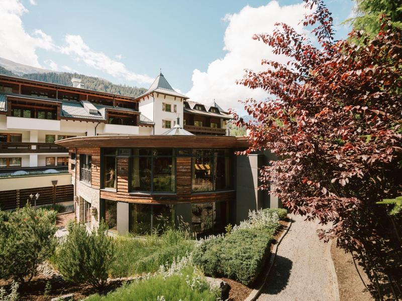 Mehrstöckiges Haus mit Holzfassade, Grünflächen, Baum mit roten Blättern und blauem Himmel.