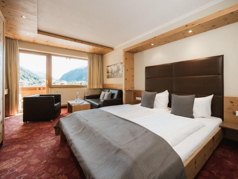 Modernes Hotelzimmer mit großem Bett, braunem Teppich, Sitzecke und Fenster mit Bergblick.