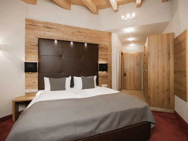 Gemütliches Schlafzimmer mit Holzbalken, braunem Kopfteil, grauer Bettdecke und warmem Licht.