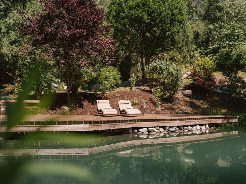 Seeufer mit zwei weißen Liegestühlen auf Holzsteg umgeben von Bäumen und grünem Wasser.