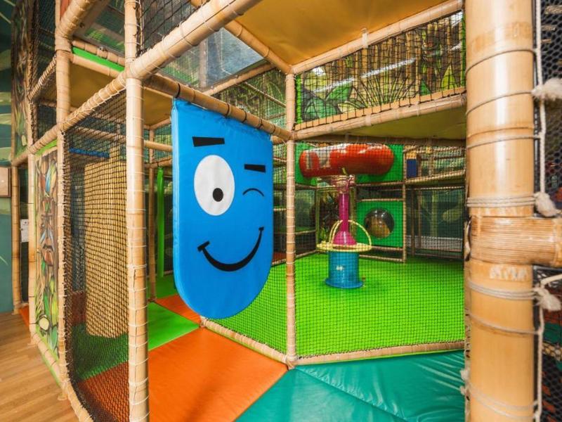 Buntes Indoor-Spielgerüst mit Kletternetz, gepolsterten Matten und fröhlichem blauen Gesicht auf Holzwänden.