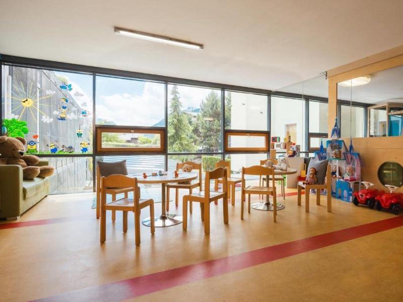 Heller, moderner Kinderspielraum mit Holzstühlen, Tischen und Spielzeugen neben großen Fenstern.