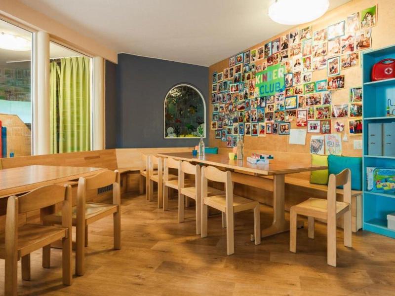 Gemütlicher Gruppenraum mit Holztischen, Stühlen und einer Fotowand in einem hellen Raum.