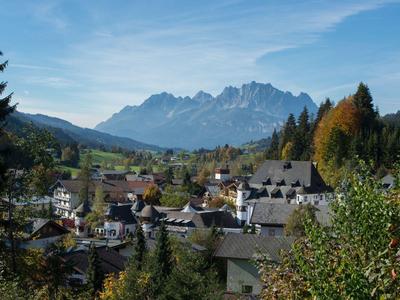 Dorf mit Häusern vor bewaldeten Hügeln und Bergen unter blauem Himmel mit wenigen Wolken.