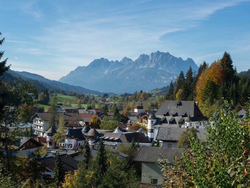 Dorf mit Häusern vor bewaldeten Hügeln und Bergen unter blauem Himmel mit wenigen Wolken.