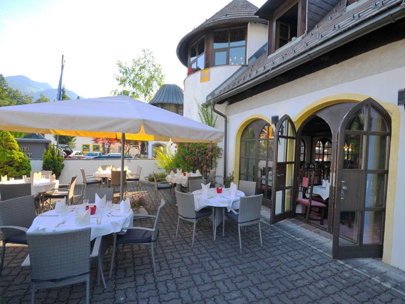 Sonnige Terrasse mit weißen Sonnenschirmen, gedeckten Tischen und Stühlen vor einem Gebäude.
