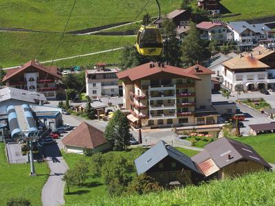 Blick auf ein Alpenhotel mit gelber Seilbahn und umliegenden Gebäuden in grüner Berglandschaft.