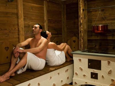 Zwei Personen sitzen entspannt in einem Holz-Saunaraum, beide in weißen Handtüchern gewickelt.