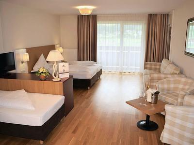 Hell eingerichtetes Zimmer mit Holzboden, zwei Betten, Sesseln, Tisch und großen Fenstern mit Gardinen.