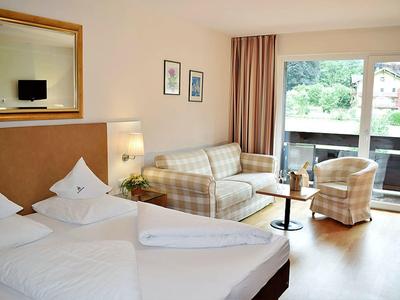 Hell eingerichtetes Hotelzimmer mit Doppelbett, Sofa, Sessel, Tisch und großem Fenster zum Balkon.