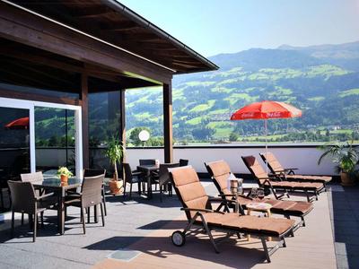 Terrasse mit Liegestühlen, Tischen und Stühlen, dahinter grüne Berge und blauer Himmel.