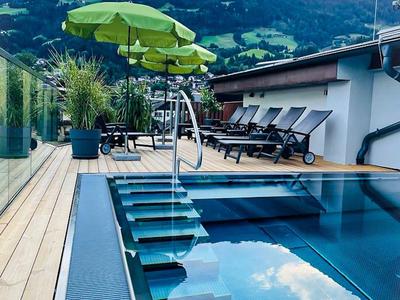 Moderner Pool mit blauem Wasser, grünen Sonnenschirmen und Liegestühlen vor Berglandschaft.