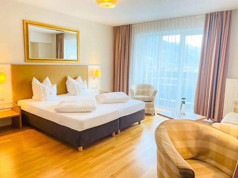 Helles Hotelzimmer mit Doppelbett, Holzfußboden, zwei Sesseln und großen Vorhängen am Fenster.