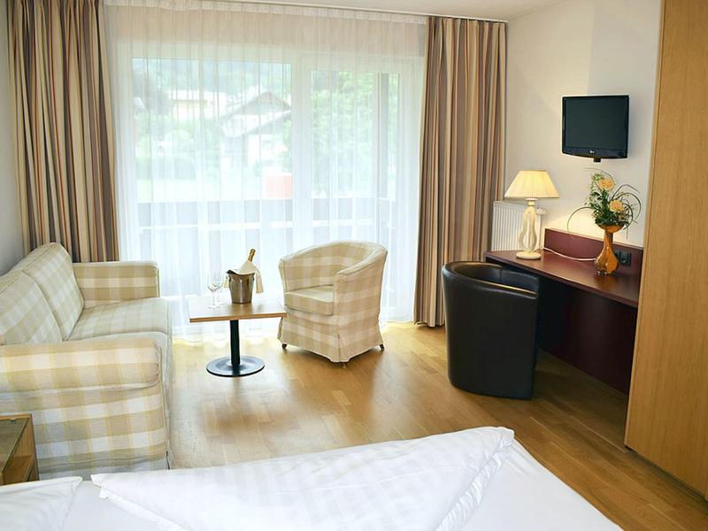 Gemütliches Hotelzimmer mit Sofa, Sessel, Tisch, Fernseher, Schreibtisch und großem Fenster mit Vorhängen.