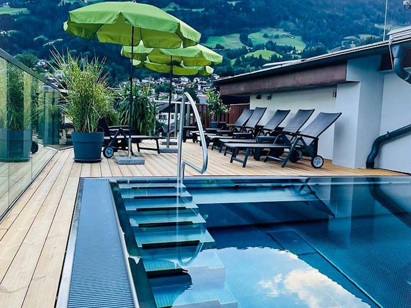 Moderner Pool mit blauem Wasser, grünen Sonnenschirmen und Liegestühlen vor Berglandschaft.