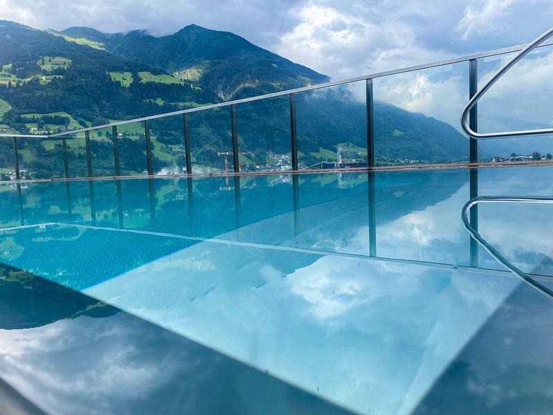 Infinity-Pool mit Glasgeländer vor Bergen und bewölktem Himmel, reflektiert das Wasser klar.